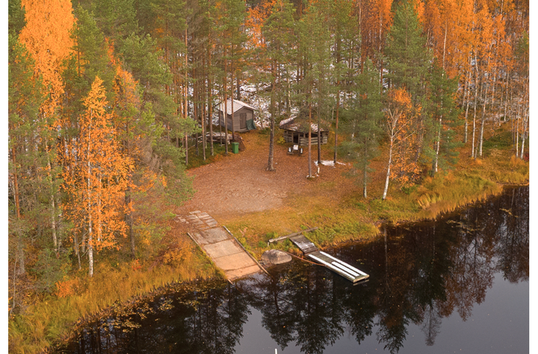 Ilmakuva tai korkealta otettu kuva syksyisest&auml; j&auml;rvimaisemasta, jossa rannalla on pieni m&ouml;kki tai leiripaikka. Ranta-alue on peitetty tummalla maalla/hiekalla ja sit&auml; ymp&auml;r&ouml;i mets&auml;, jonka puissa on voimakkaat keltaiset ja oranssit ruskan v&auml;rit. Kuvassa n&auml;kyy kaksi erilaista, puista laituria ulottumassa tummaan veteen: toinen leve&auml;mpi ja lyhyempi, toinen kapeampi ja pidempi.