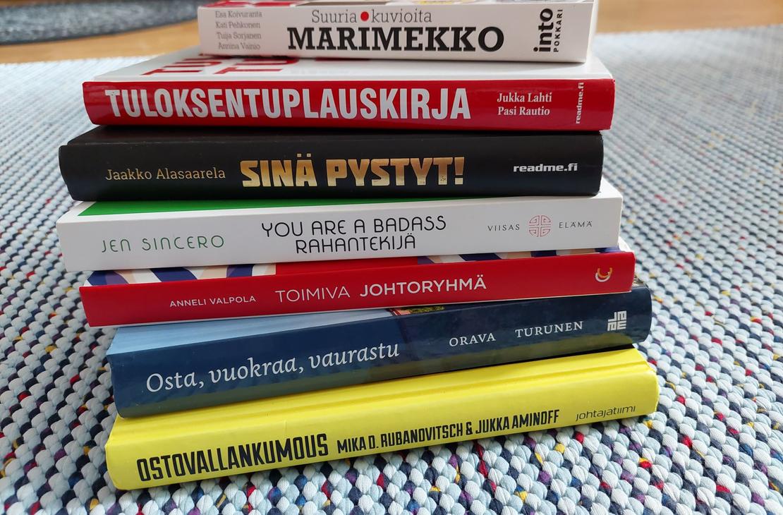 kuvassa on pino kirjoja, jotka on pinottu päällekkäin. Kirjojen kannet ovat erivärisiä ja -kuvioisia, ja niissä on erilaisia tekstejä. Pinon päällimmäinen kirja on vaaleansininen ja siinä lukee "MARIMEKKO". Pinon alimmainen kirja on keltainen ja siinä lukee "OSTA VAIN, JOS TARVITSET". Muissa kirjoissa on tekstejä kuten "TULOKSENTUPLAUSKIRJA", "SINÄ PYSTYT!", "YOU ARE A BADASS", "JOHTAJA ILMAN JOUKKOJA", "OMA RAHA, OMAT SÄÄNNÖT". Kirjat on asetettu harmaalle matolle.