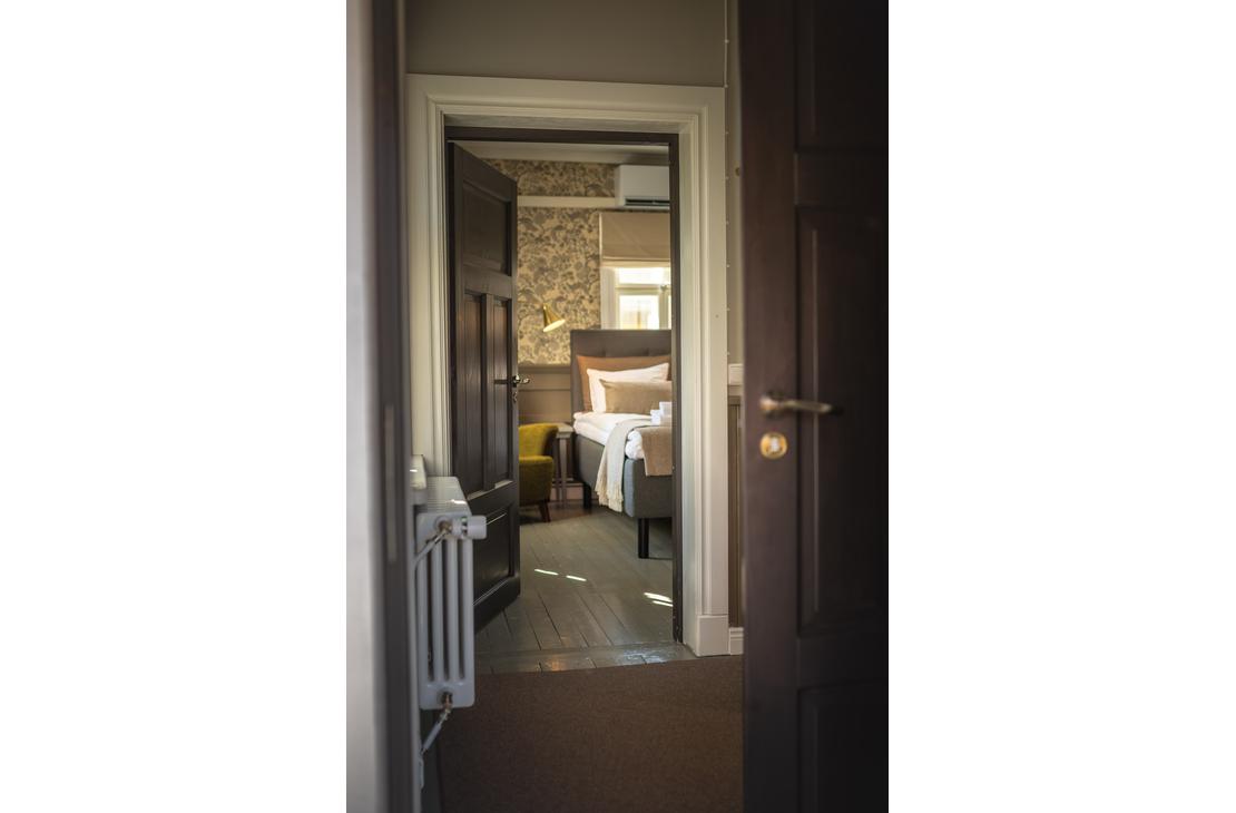 Kuva on otettu Matkailukeskus Puu-Juuka Oy:n huoneen ovelta, ja se esittää auki olevaa, tummaa puuovea, josta avautuu näkymä hotellihuoneeseen. Etualalla vasemmalla näkyy vanhanaikainen valkoinen patteri. Huoneessa, jonne ovi johtaa, on leveä sänky neutraalin sävyisillä petivaatteilla, limevihreä nojatuoli ja tumma, kuvioitu tapetti. Lattia huoneiden välillä on tummaa puuta.