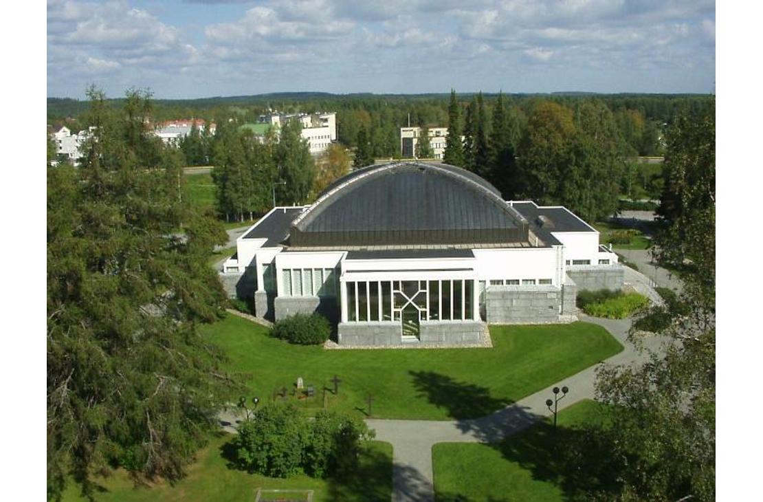 Ilmakuva Lieksan kirkosta kesällä. Valkoisessa rakennuksessa on suuri, kupolimainen tumma katto, ja sitä ympäröi vihreä nurmikko ja puusto.