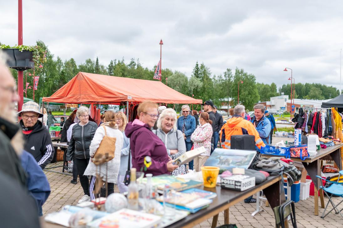 Ihmisiä tutkimassa tavaroita Ilomantsin ulkoilmatorin myyntipöydillä pilvisenä päivänä. Etualalla on pöytä täynnä pientavaraa, kuten kirjoja ja laatikoita, ja taustalla näkyy suuri oranssi myyntiteltta sekä vaaterekkejä.