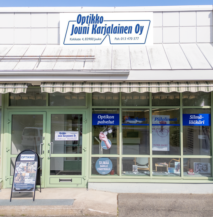 Kuva Optikko Jouni Karjalainen Oy -liikkeen ulkopuolesta aurinkoisena päivänä. Liikkeessä on suuret ikkunat ja vaaleanvihreä ovi. Ikkunoissa on mainosteippauksia, joissa mainitaan "Optikon palvelut", "Piilolinssit Kuno-Qvist" ja "Silmälääkäri". Sisäänkäynnin yläpuolella on markiisi. Rakennuksen yläosassa on suuri, kaarevalla kehyksellä varustettu kyltti, jossa lukee "Optikko Jouni Karjalainen Oy", sekä osoite "Kirkkotie 4, 83900 Juuka" ja puhelinnumero "Puh. 013 470 577".