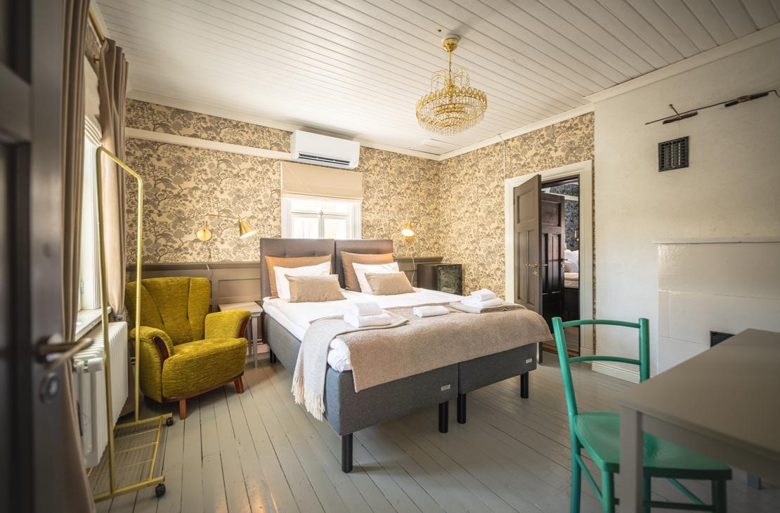 Kuva esittää Matkailukeskus Puu-Juuka Oy:n hotellihuonetta tai majoitustilaa. Huoneessa on leveä, kaksiosainen parisänky neutraalin sävyisillä petivaatteilla. Sängyn takana seinällä on karkea, kivinen tai puinen tapetti. Huoneessa on vaaleansininen puulattia, ja vasemmalla on sammaleenvihreä nojatuoli. Oikealla on pieni pöytä ja kaksi turkoosinsinistä tuolia. Huone on valoisa ja siinä on moderni sisustus.