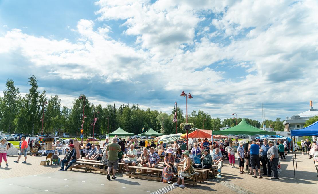 Ihmisiä istumassa puupenkeillä ja kiertelemässä Ilomantsin torilla värikkäiden myyntitelttojen välissä aurinkoisena päivänä suuren, valkopilvisen taivaan alla.