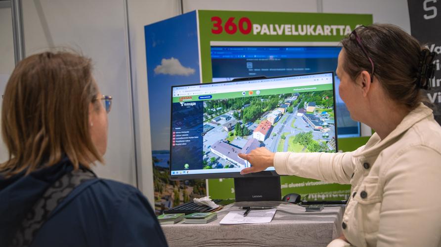 Kaksi naista messuosastolla. Toinen nainen osoittaa sormella tietokoneen näyttöä, jolla näkyy ilmakuva 360 Palvelukartasta. Taustalla on kyltti, jossa lukee "360 PALVELUKARTTA".