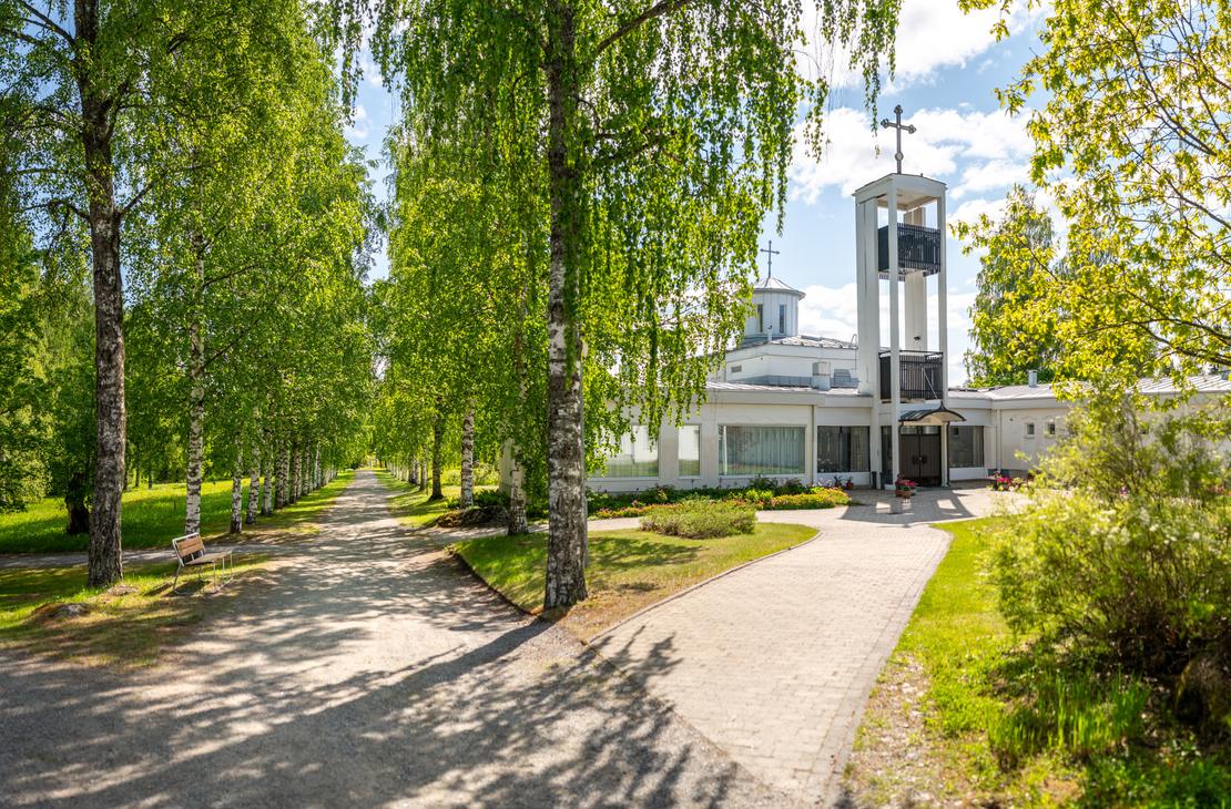 Laajakulmakuva Lintulan luostarin sisäänkäynnille aurinkoisena päivänä. Vasemmalla reunustaa vehreä koivukuja, ja oikealla laatoitettu käytävä johtaa moderniin, valkoiseen luostarirakennukseen, jossa on erillinen kellotapuli ja risti.