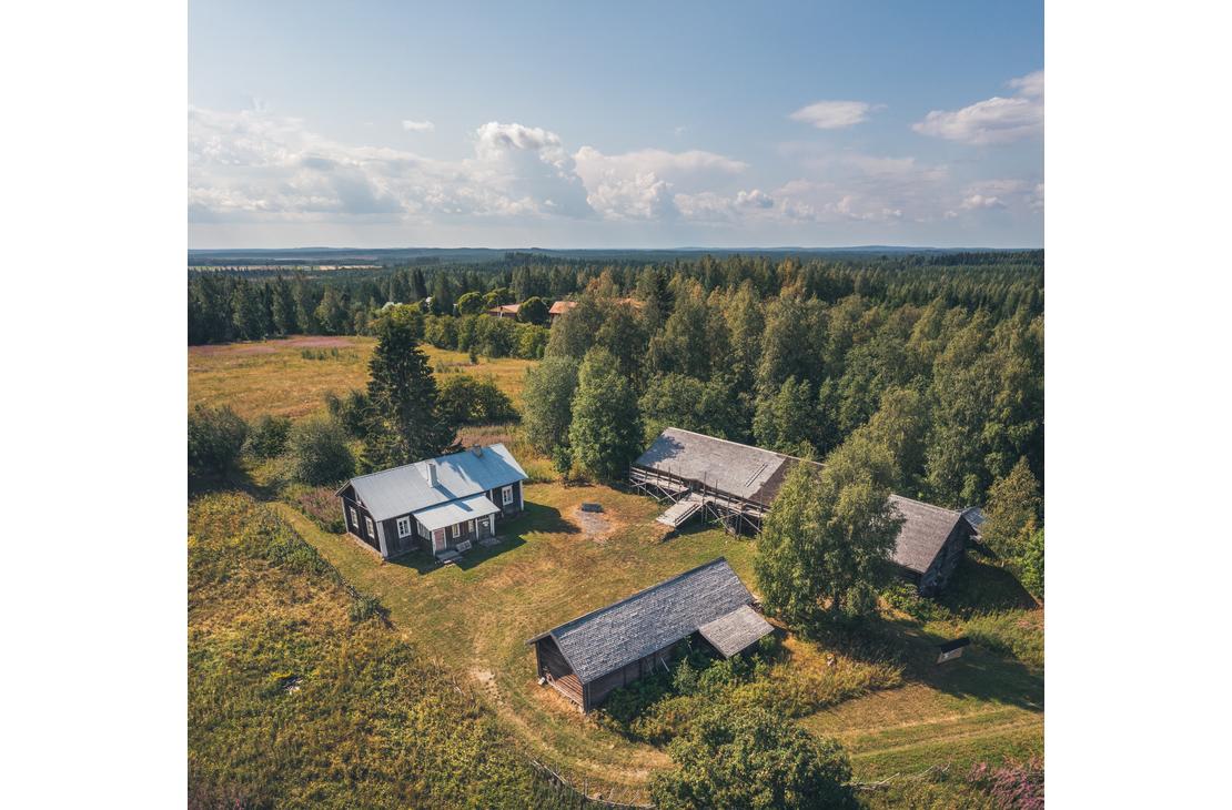 Ilmakuva Makkolan museotilasta, joka koostuu useista perinteisistä, tummista hirsirakennuksista harmailla katoilla. Rakennukset sijaitsevat vihreällä niityllä, jota ympäröi tiheä metsä ja aukea pelto. Piha-alueella näkyy ajopolkuja ja pieni avoin alue rakennusten välissä.