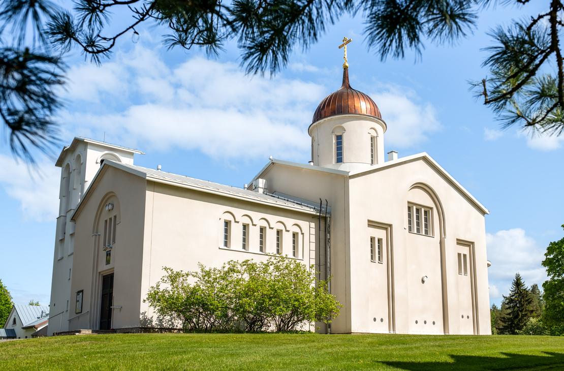 Valamon luostarin vaalea kirkkorakennus, jossa on kuparinen sipulikupoli ja ortodoksinen risti, kuvattuna alaviistosta aurinkoisena p&auml;iv&auml;n&auml;. Etualalla on vihre&auml; nurmikko, ja kuvan yl&auml;osaa kehyst&auml;v&auml;t m&auml;nnynoksat.