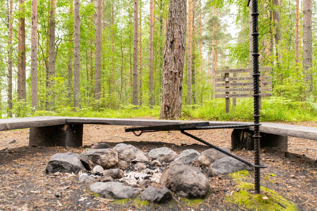 Lähikuva maastossa olevasta nuotiopaikasta ja grilliritilästä. Taustalla näkyy puinen opasteviitta, jossa on reittien ja lampien nimiä, kuten Hiislampi ja Taitajan taival.