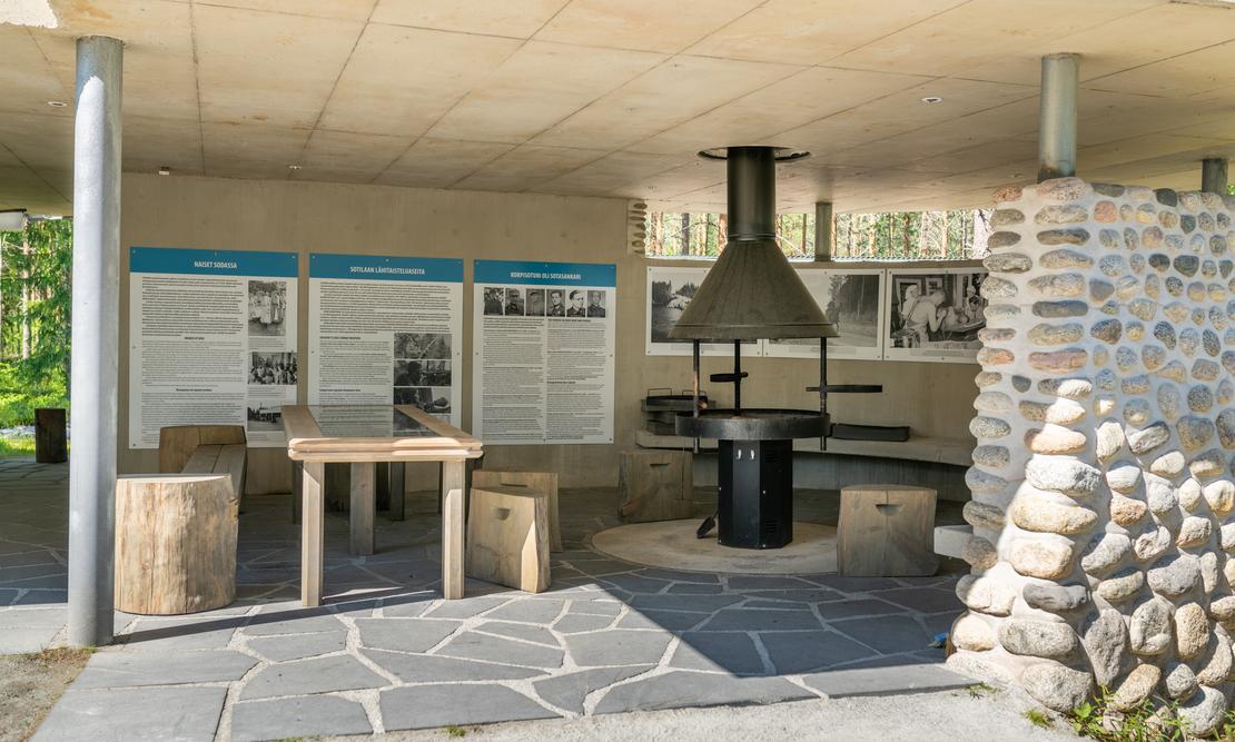 Sisäkuva Öykkösenvaaran taistelualueen taukopaikasta/museorakennuksesta. Kuvassa näkyy moderni, kivetty lattia, jolla on grillauspaikka tai takka suuren mustan liesituulettimen alla. Seinällä on kolme infotaulua teksteineen ja mustavalkokuvineen. Edessä on yksinkertainen puinen pöytä ja istuimina toimivia puunrunkoja. Oikealla näkyy luonnonkivillä verhoiltu pilarin osa. Tila on valoisa ja katettu.