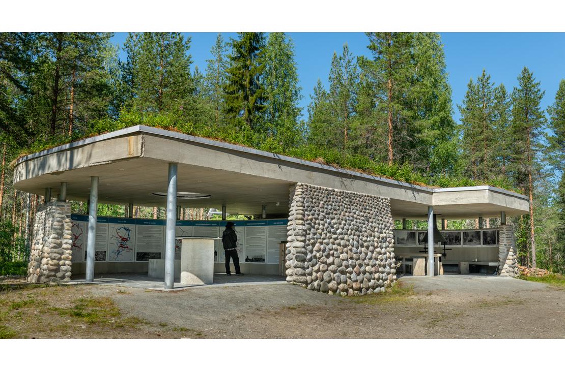 Kuva &Ouml;ykk&ouml;senvaaran taistelualueen taukopaikasta tai museorakennuksesta. Se on moderni, matala, osittain avoin katos, jossa on vihre&auml;, maanpeitt&auml;m&auml; katto. Rakenne koostuu betonista ja py&ouml;reill&auml; luonnonkivill&auml; verhoilluista pylv&auml;ist&auml;. Rakennuksen alla on taukopaikka tai n&auml;yttelytila. Rakennus sijaitsee mets&auml;n reunassa soraisella maapohjalla.