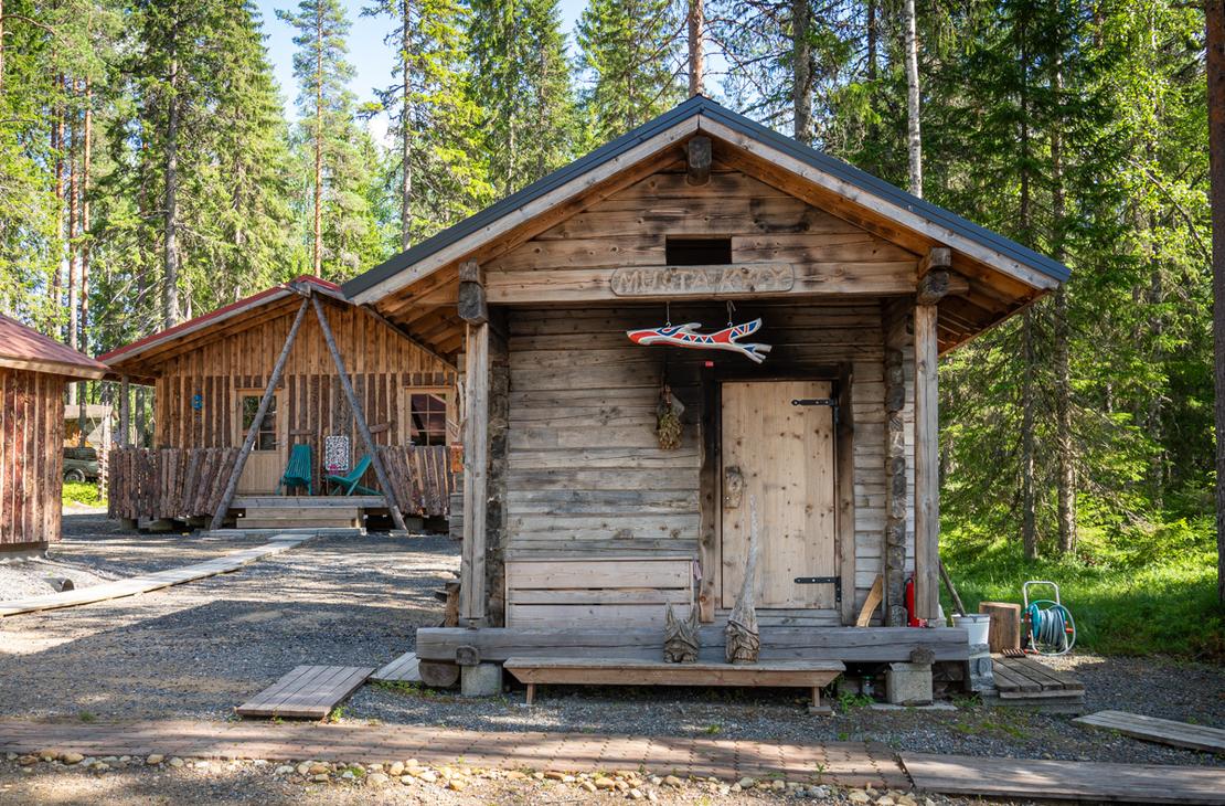 Harmaantuneesta hirrestä rakennettu perinteinen sauna metsän siimeksessä. Oven yläpuolella on puinen kyltti, jossa lukee "MUSTA KYPY", ja terassilla on puisia koristeveistoksia.
