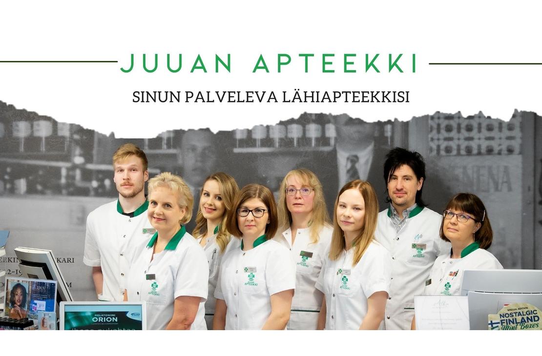 Ryhmäkuva Juuan Apteekin henkilökunnasta, jossa seitsemän naista ja kaksi miestä seisovat rivissä valkoisissa takeissa. Naiset etualalla ovat pukeutuneet valkoisiin takkeihin, joissa on vihreät kaulukset ja taskut. Taustalla on mustavalkoinen kuva tiilitalosta tai historiallisesta rakennuksesta. Kuvan yläosassa lukee vihreällä tekstillä "JUAN APTEEKKI" ja alapuolella pienemmällä tekstillä "SINUN PALVELEVA LÄHIAPTEEKKISI".