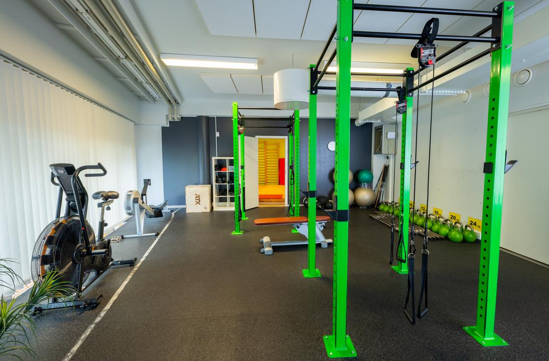 Kuva esittää Kinesiocenter Kuntoilukeskuksen harjoitustilaa. Lattiana on tumma kuminen lattia, ja tilassa on kirkkaan limevihreitä metallisia telineitä crossfit-tyyliseen harjoitteluun. Vasemmalla näkyy kuntopyörä tai ergometri. Oikealla taustalla on rivi kahvakuulia ja keskellä näkyy avoimen oven kautta seuraava huone, joka on keltainen ja punainen.
