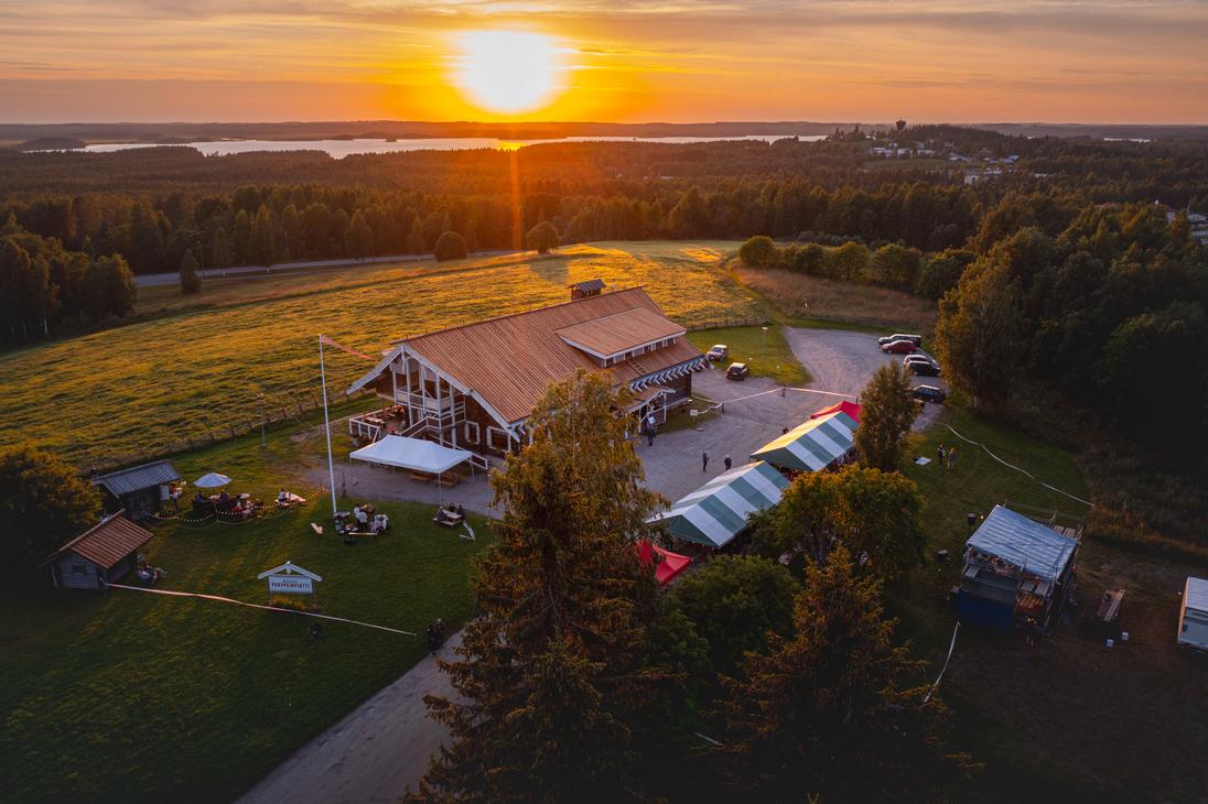 Ilmakuva Parppeinpirtistä ja sitä ympäröivästä metsä- ja järvimaisemasta kirkkaassa auringonlaskussa. Laskeva aurinko heijastaa kultaista valoa koko näkymään.