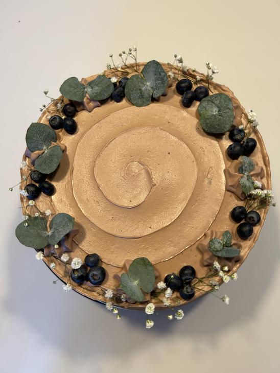 Yläviistosta otettu kuva vaaleanruskealla kuorrutteella päällystetystä kakusta, joka on koristeltu reunoilta mustikoilla, pienillä valkoisilla kukilla ja eukalyptuksen lehdillä.