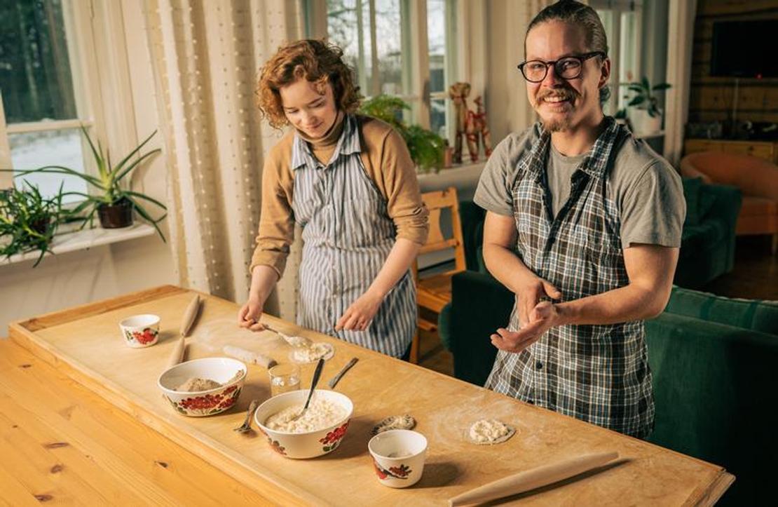 Sisäkuva, jossa kaksi aikuista, nainen ja mies, leipovat perinteisiä karjalanpiirakoita puisen pöydän ääressä. Molemmat henkilöt ovat pukeutuneet essuihin. Mies oikealla katsoo suoraan kameraan ja hymyilee. Pöydällä on taikinakulhoja ja valmiita, ohuita taikinapohjia. Tila on lämminhenkinen, ja taustalla näkyy ikkuna ja kasveja. Toiminta liittyy todennäköisesti Äksyt Ämmät -yrityksen karjalanpiirakka-työpajoihin tai -palveluihin.