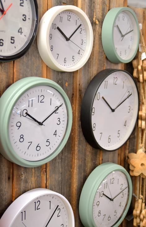 Ryhmä pyöreitä seinäkelloja, joiden kehykset ovat eri värisiä, kuten mintunvihreitä, valkoisia ja mustia, kiinnitettynä rouheaan puuseinään.