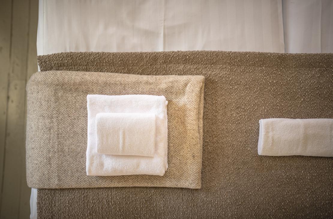 Lähikuva hotellihuoneen sängyn jalkopäästä tai päädystä Matkailukeskus Puu-Juuka Oy:ssä. Sänky on pedattu valkoisilla lakanoilla. Patjan päällä on ruskea sängynpeite tai leveä päiväpeite. Peitteen päällä on siististi taiteltu kasa valkoisia pyyhkeitä, ja oikealla on yksi erillinen pyyhe.