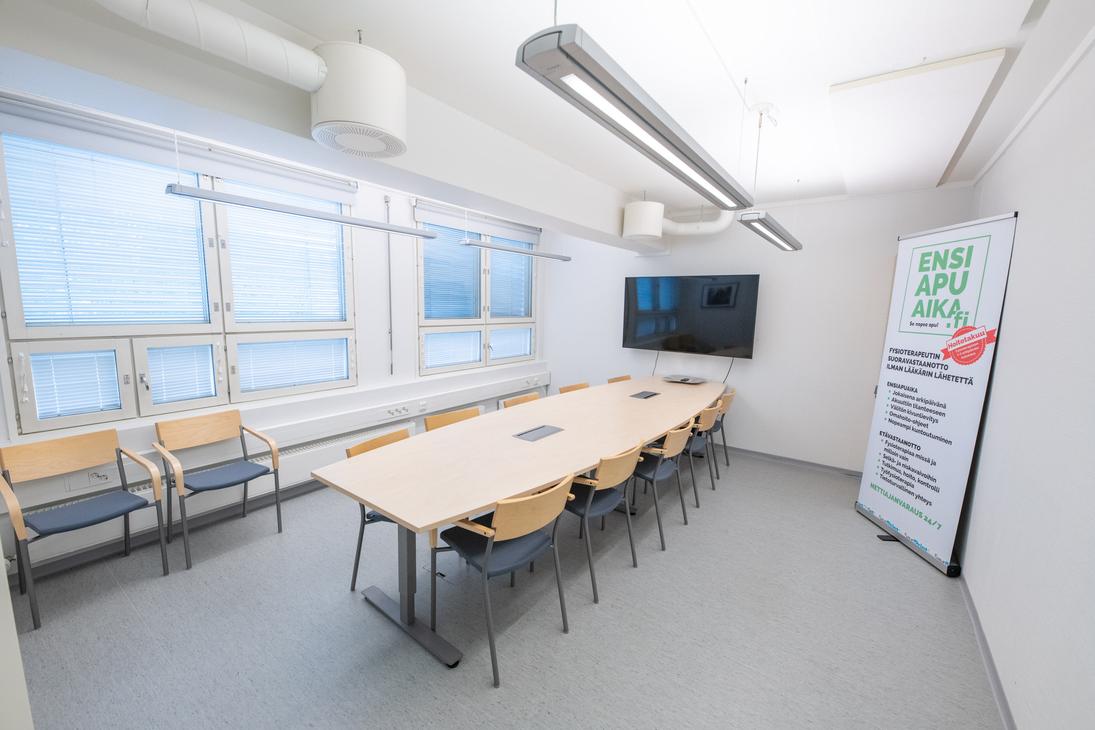 Kuva esittää Liikekulman Toimistohotellin isoa, valoisaa neuvottelutilaa tai koulutushuonetta. Huoneessa on vaalea lattia, ja keskellä on suuri, vaaleapuinen neuvottelupöytä, jonka ympärillä on mustia tuoleja. Pöydän päällä on liitäntöjä. Oikealla seinällä on suuri taulutelevisio ja pystyssä oleva roll-up-mainos. Vasemmalla on pitkä ikkunanauha, jossa on siniset sälekaihtimet, ja seinää vasten on muutama ylimääräinen vierastuoli.