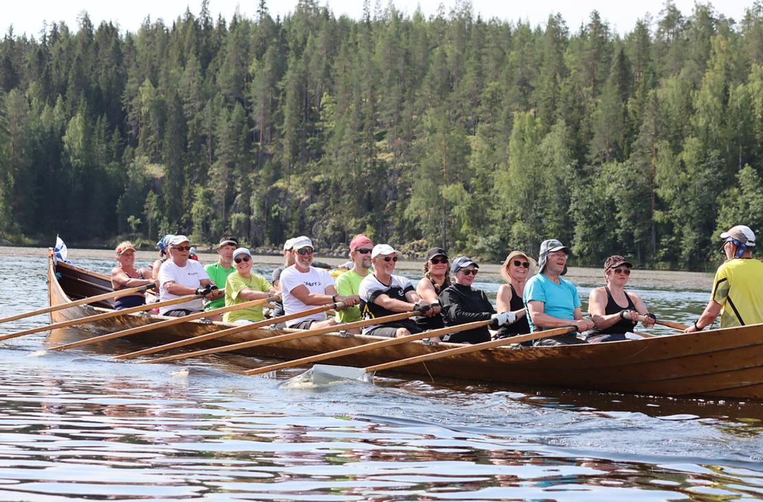 Pitk&auml; puinen kirkkovene t&auml;ynn&auml; soutajia lipuu tyynell&auml; j&auml;rvell&auml;. Taustalla n&auml;kyy jyrkk&auml; mets&auml;inen rinnerynn&auml;s.