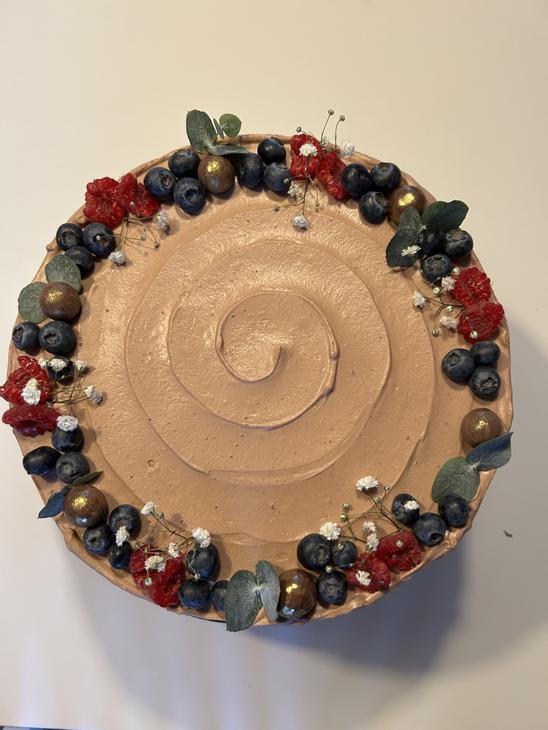 Pyöreä kakku, jonka reunoilla on runsas koristeena mustikoita, tummanpunaisia kukkia, vihreitä lehtiä ja kultaisia koristekuulia.