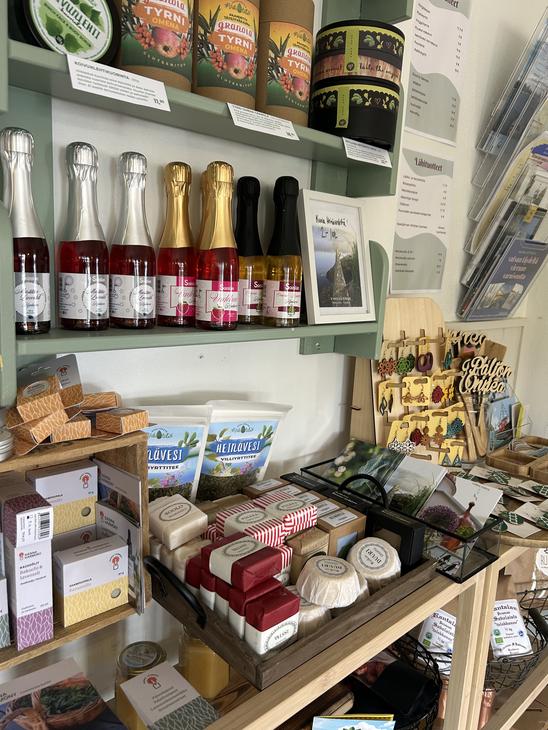 Lähikuva Satamakahvila-ravintola Kermacotan myymälähyllystä, jossa on tarjolla erilaisia paikallisia tuotteita ja matkamuistoja. Hyllyillä näkyy muun muassa pieniä kuohuviinipulloja, hilloja, paikallisia elintarvikkeita sekä käsitöitä. Tuotteet on aseteltu siististi puisiin ja vaaleanvihreisiin hyllykköihin.