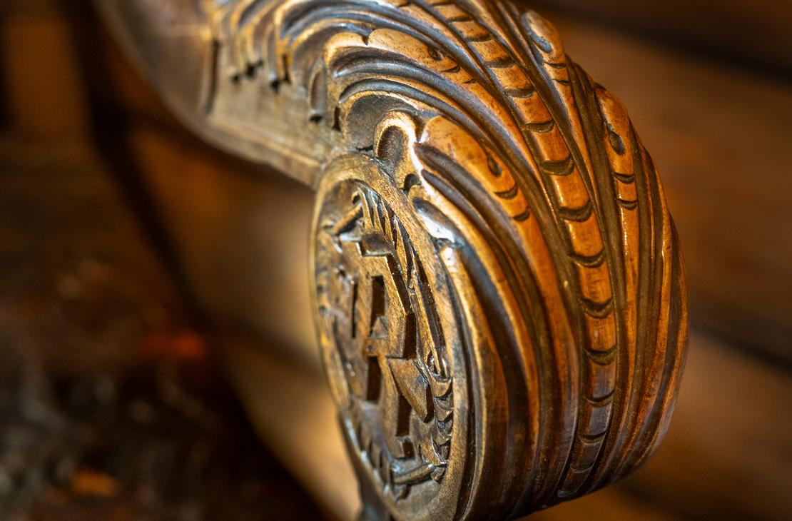 Lähikuva tumman puisen kaiteen tai tuolin käsinojan päädystä, jossa on taidokkaasti veistetty, koristeellinen kuvio. Kaiverrus on pyöreä ja kupera, muistuttaen simpukankuorta tai sulkakuviointia, ja sen keskellä on erottuva, geometrinen kuvio. Käsinoja on kiillotettu ja kiinnittyy tummaan pylvääseen. Tausta on epätarkka, mutta siinä näkyy puupintoja.