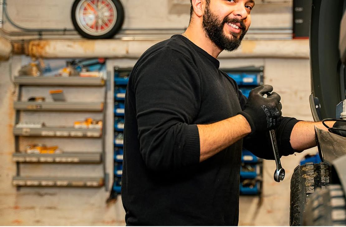 Parrakas mekaanikko hymyilee kameralle kiintoavain kädessään korjaamolla ajoneuvon äärellä. Kuvan alalaidassa on Lieksan Koneasema -tekstilogo.