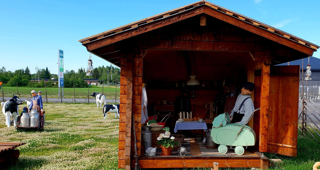 Kuva esittää Juuan Apteekin perinnepihaa aurinkoisena päivänä. Etualalla on pieni, puinen katos tai koju tummasta puusta, jossa on näytteillä vanhanaikaisia esineitä, kuten nukke tai patsas perinteisissä vaatteissa keinutuolissa sekä pöytäliinoja ja kukkia. Vasemmalla nurmialueella näkyy ihmisiä ja maalattuja lehmäpatsaita tai koristeita. Taustalla erottuu lipputanko ja kadunvarsirakennuksia.
