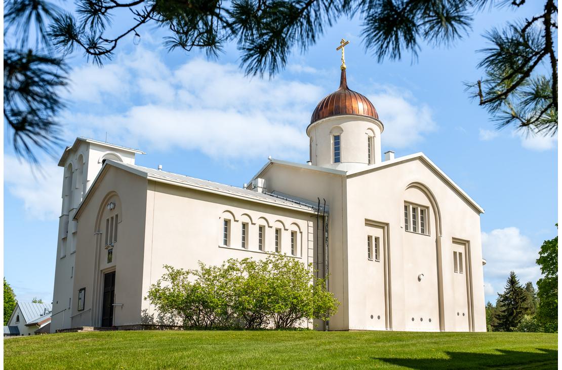 Valamon luostarin vaalea kirkkorakennus, jossa on kuparinen sipulikupoli ja ortodoksinen risti, kuvattuna alaviistosta aurinkoisena päivänä. Etualalla on vihreä nurmikko, ja kuvan yläosaa kehystävät männynoksat.