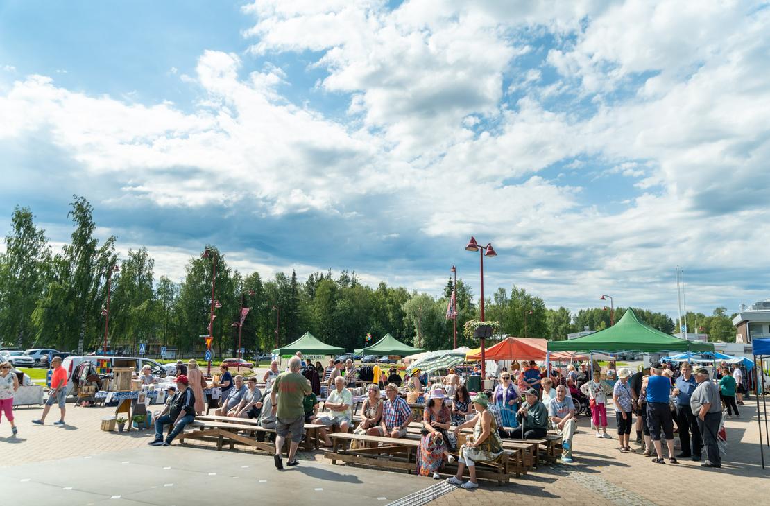 Ihmisiä istumassa puupenkeillä ja kiertelemässä Ilomantsin torilla värikkäiden myyntitelttojen välissä aurinkoisena päivänä suuren, valkopilvisen taivaan alla.