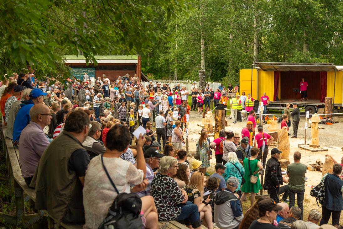 Laaja kuva ulkoilmatapahtumasta, luultavasti Karhufestivaaleilta, jossa suuri joukko ihmisiä on kokoontunut katsomaan esitystä tai kilpailua. Etualalla osa yleisöstä istuu puisilla katsomoilla, ja taka-alalla seisoo lisää yleisöä. Oikealla on keltainen lava tai esiintymislava, jolla on esiintyjä. Lavan edessä on työstettyjä puuveistoksia ja työntekijöitä keltaisissa liiveissä. Taustalla näkyy metsän reuna ja pieni, punainen rakennus.