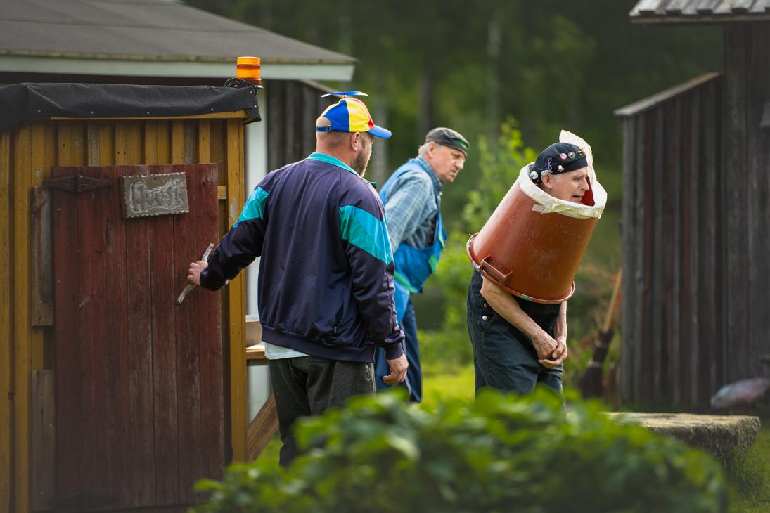 Möhkön kesäteatterin Peitetarina-näytelmän kohtaus, jossa kolme miestä esiintyy ulkonäyttämöllä. Vasemmalla mies, jolla on värikäs potkurilippis, on avaamassa puisen ulkokäymälän ovea, jossa on teksti "Huussi". Oikealla vanhempi mies seisoo vartalonsa ympärille asetetun suuren ruskean muovisaavin sisällä ilmehtien vaivautuneesti, ja heidän takanaan kolmas mies seuraa tilannetta.