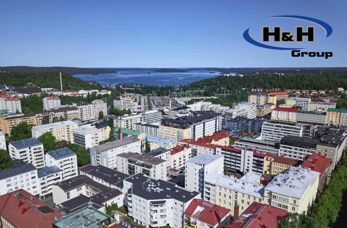 Ilmakuva kaupungin keskustasta, jossa näkyy tiivistä rakentamista ja asuinkerrostaloja. Taustalla siintää järvimaisema ja metsää, ja oikeassa yläkulmassa on H&H Groupin logo.
