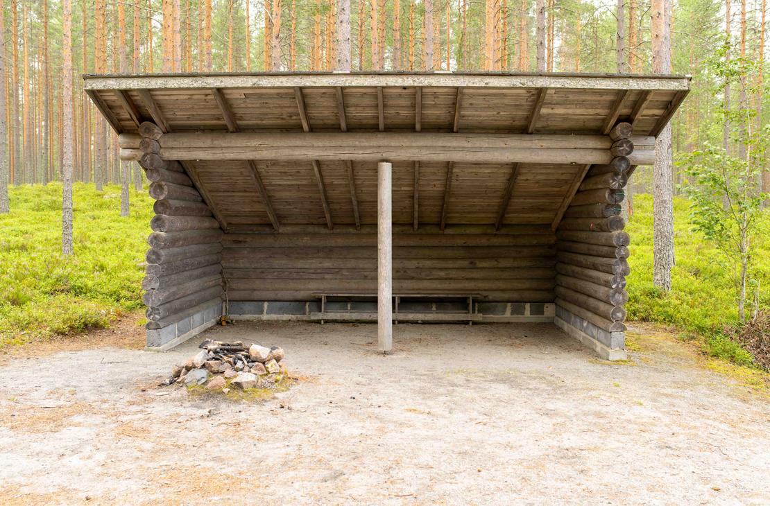 Suuri hirsirakenteinen laavu mäntymetsän siimeksessä. Laavun edustalla hiekalla on kivistä koottu nuotiopaikka ja sisällä pystytuki sekä puupenkki.
