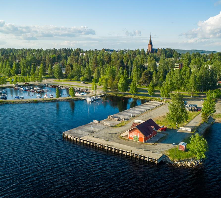 Ilmakuva Nurmeksen satamasta Pielisen rannalla aurinkoisena päivänä. Etualalla on laituri ja punainen satamarakennus, taustalla venesatama ja vehreän metsän yläpuolelle kohoava Nurmeksen kirkon torni.