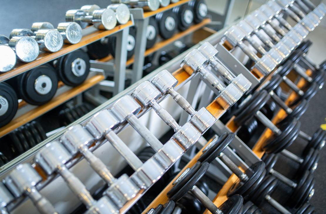 Lähikuva Kinesiocenter Kuntoilukeskuksen kuntosalin käsipainotelineestä. Telineessä on riveittäin hopeanvärisiä, metallisia käsipainoja eri kokoisina, alkaen pienemmistä (etualla) ja jatkuen suurempiin (taustalla). Käsipainot on aseteltu viistosti telineisiin, joissa on oranssit tai keltaiset hyllyt.