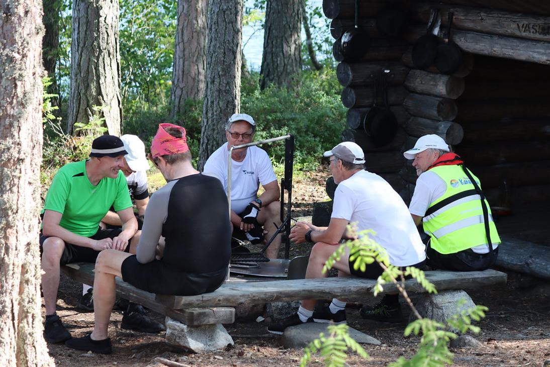 Ryhm&auml; miehi&auml; istuu puupenkeill&auml; nuotiopaikan &auml;&auml;rell&auml; mets&auml;ss&auml; sijaitsevan hirsilaavun edustalla.