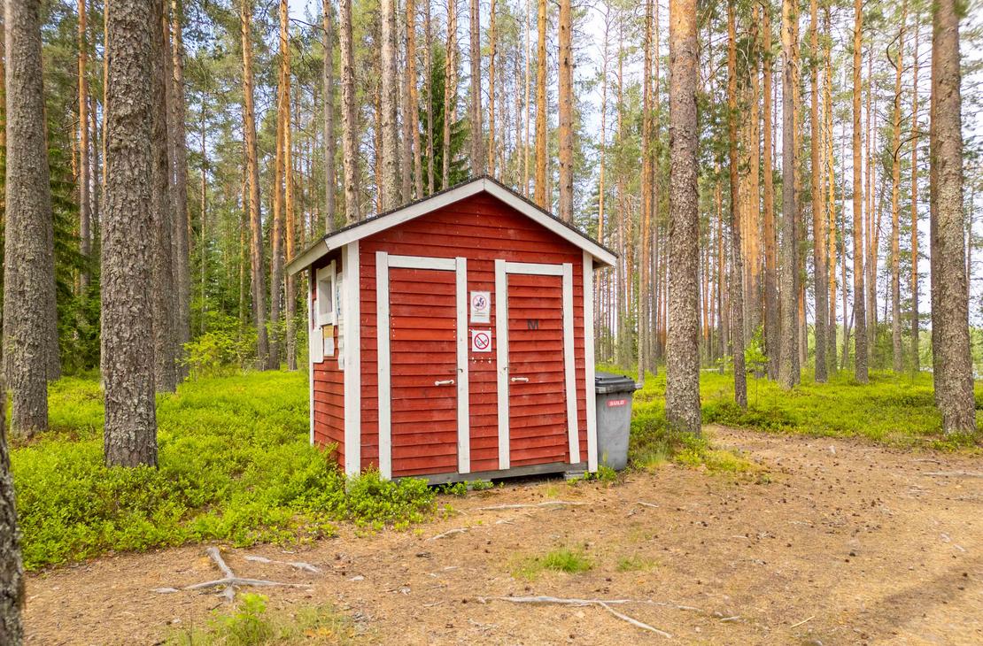 Punainen valkonurkkainen kuivakäymälä rakennus mäntymetsän keskellä. Rakennuksen vieressä on harmaa roska-astia ja maasto on käpystä kangasmaata.
