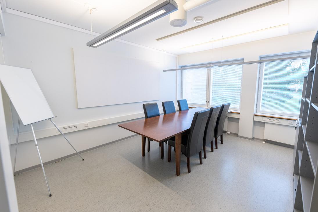 Kuva esittää Liikekulman Toimistohotellin pientä neuvotteluhuonetta. Huoneessa on vaaleanharmaa lattia ja valkoiset seinät. Keskellä on tummanruskea neuvottelupöytä ja sen ympärillä mustat nahka- tai kangasverhoillut tuolit. Vasemmalla on valkoinen fläppitaulu ja suuri valkoinen seinätaulu/valkokangas. Ikkunoista näkyy ulos vihreää. Oikealla näkyy harmaa hyllykkö.