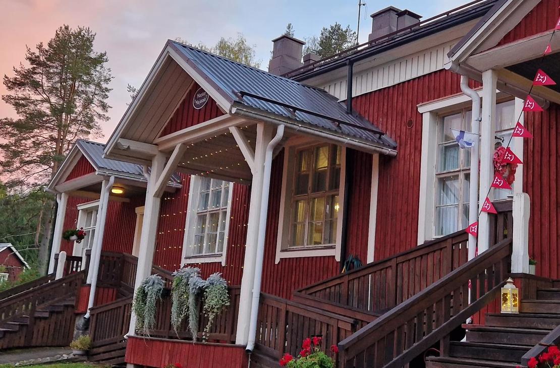 Lähikuva Mantan Majatalon sisäänkäynnistä. Punamultaisessa puutalossa on puiset portaat ja katettu kuisti, jossa on valkoiset pylväät ja kaiteet. Kuistia on koristeltu punaisilla kukilla tai nauhoilla. Sisäänkäynti on keltainen. Kuva on otettu matalalta kulmalta, ja taustalla näkyy metsän reunaa. Valo luo pitkiä varjoja, viitaten auringonlaskuun tai -nousuun.