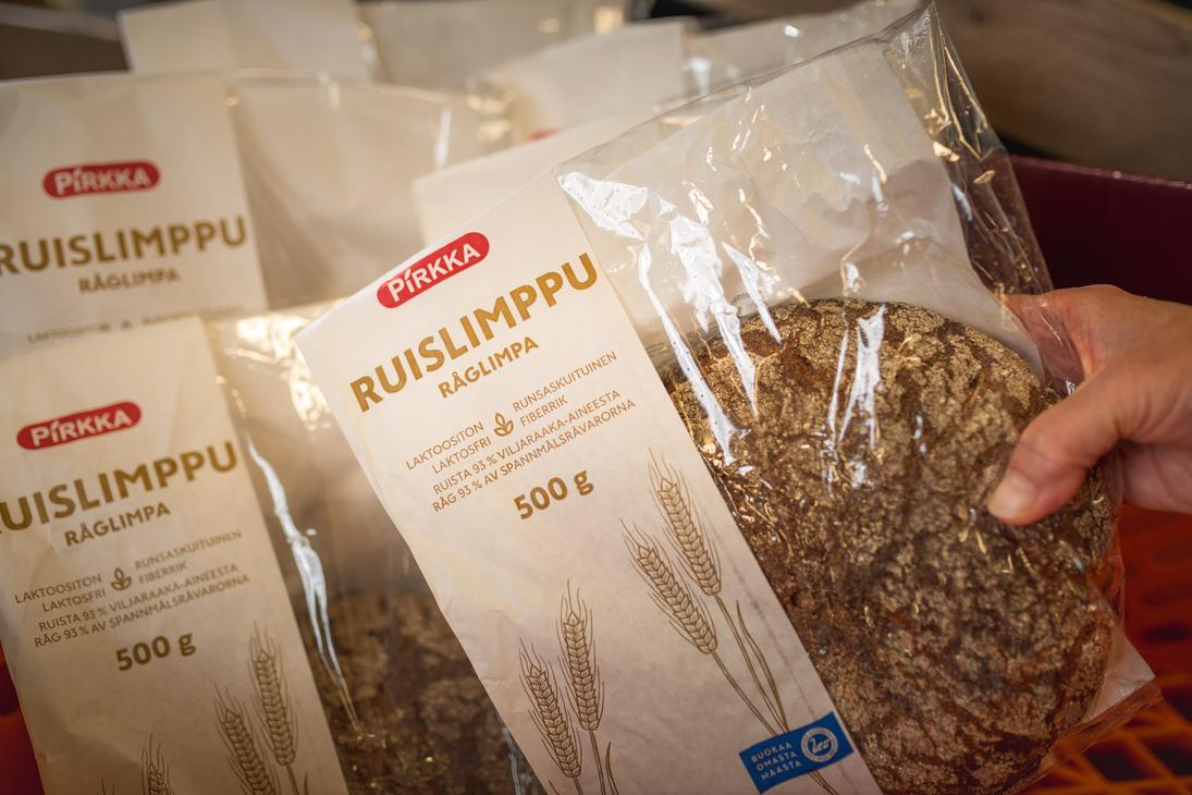 Käsi pitelee osittain läpinäkyvää Pirkka Ruislimppu -leipäpakkausta K-Market Ilomantsissa. 500 gramman pakkauksesta erottuu pyöreä ruisleipä sekä tekstit ”Pirkka Ruislimppu Råglimpa”. Taustalla on useita muita samanlaisia leipäpakkauksia rivissä.