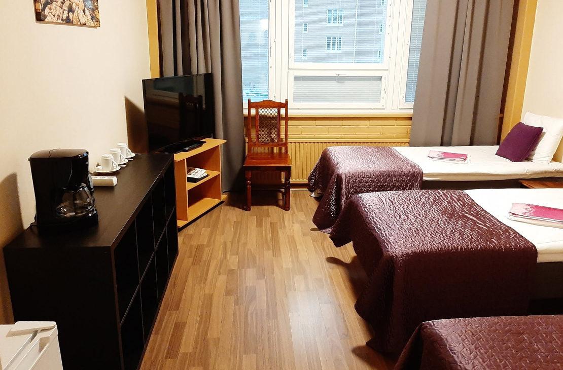 Hotellihuone, jossa on kolme sänkyä tummanpuhu villasävyisillä päiväpeitoilla. Huoneessa on puulaminaattilattia, televisio, tumma lipasto kahvinkeittimineen sekä ikkuna, jossa on harmaat verhot.