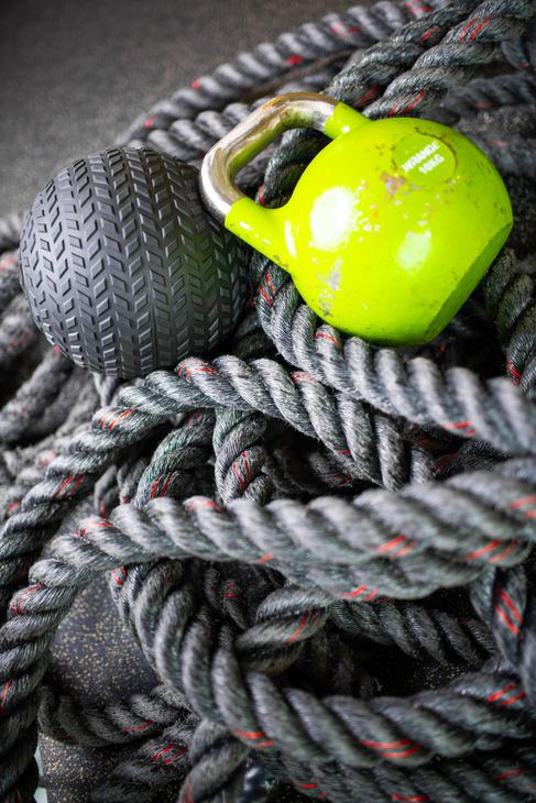 Lähikuva Kinesiocenter Kuntoilukeskuksen kuntosalilaitteista. Köysien ja painojen kasauma on etualalla: tummanharmaa harjoitusköysi (taisteluköysi), kirkkaan limevihreä kahvakuula ja tumma, kuvioitu painopallo (medicine ball). Kuva symboloi tehokasta ja monipuolista harjoittelua.