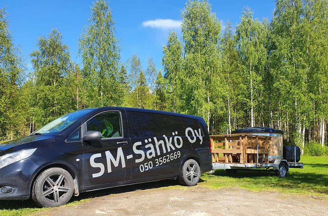 Kuva esittää SM-Sähkö Oy:n mustaa pakettiautoa, jonka kyljessä lukee valkoisella, isolla tekstillä "SM-Sähkö Oy" ja alapuolella puhelinnumero "050 3562669". Auto on pysäköity nurmialueelle, ja se vetää perässään peräkärryä, jossa on puinen, pyöreä palju tai ulkoporeamme. Taustalla on tiheä, vehreä koivumetsä aurinkoisena kesäpäivänä kirkkaansinisen taivaan alla.