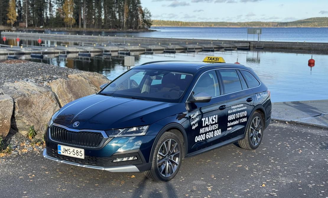 Kuva esittää tummansinistä, farmarimallista taksia (Skoda Octavia) pysäköitynä satama-alueella. Auton katolla on keltainen "TAKSI" -kyltti. Kyljessä on useita tekstejä, joista erottuvat selkeästi sanat "TAKSI HEINÄVESI 0400 600 800" valkoisella, sekä puhelinnumeroita. Auto on pysäköity sora-alueelle suuren kiven viereen. Taustalla näkyy tyyni järvimaisema ja venelaitureita.