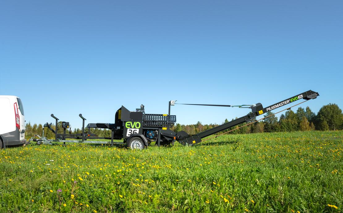 KLAPikosen musta ja vihreä Pilkemaster EVO 36 -klapikone seisoo aurinkoisella, keltaisten voikukkien täplittämällä niityllä valkoisen pakettiauton perässä, kirkkaansinisen taivaan alla.