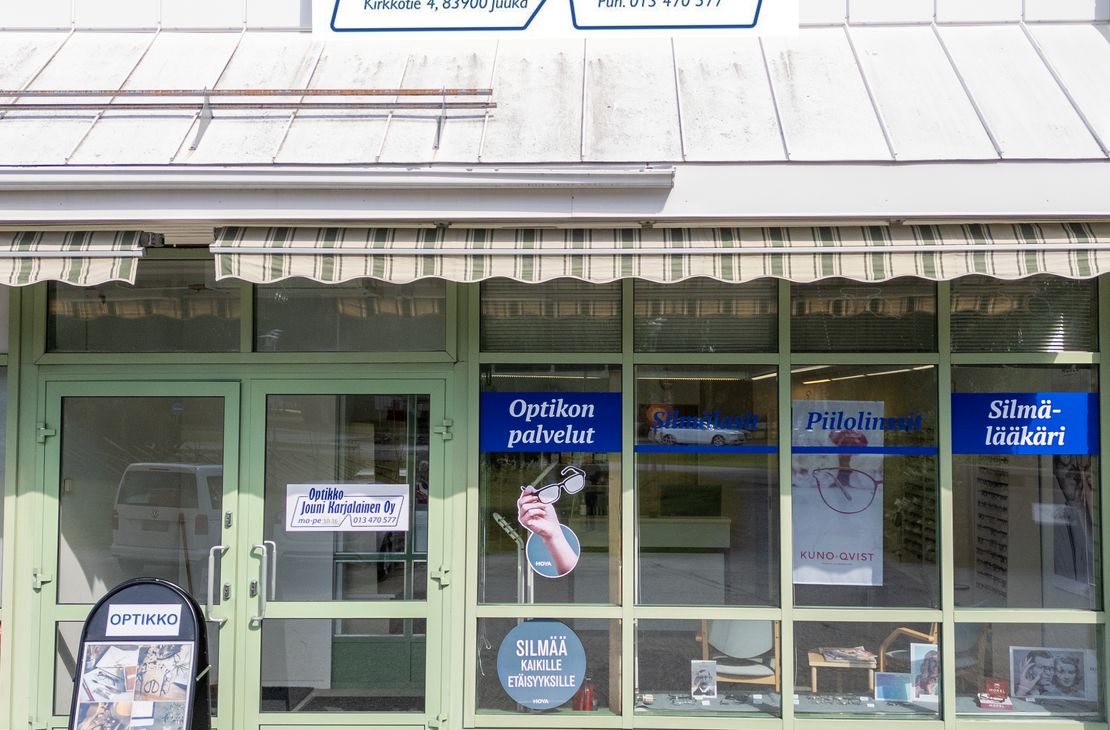 Kuva Optikko Jouni Karjalainen Oy -liikkeen ulkopuolesta aurinkoisena päivänä. Liikkeessä on suuret ikkunat ja vaaleanvihreä ovi. Ikkunoissa on mainosteippauksia, joissa mainitaan "Optikon palvelut", "Piilolinssit Kuno-Qvist" ja "Silmälääkäri". Sisäänkäynnin yläpuolella on markiisi. Rakennuksen yläosassa on suuri, kaarevalla kehyksellä varustettu kyltti, jossa lukee "Optikko Jouni Karjalainen Oy", sekä osoite "Kirkkotie 4, 83900 Juuka" ja puhelinnumero "Puh. 013 470 577".