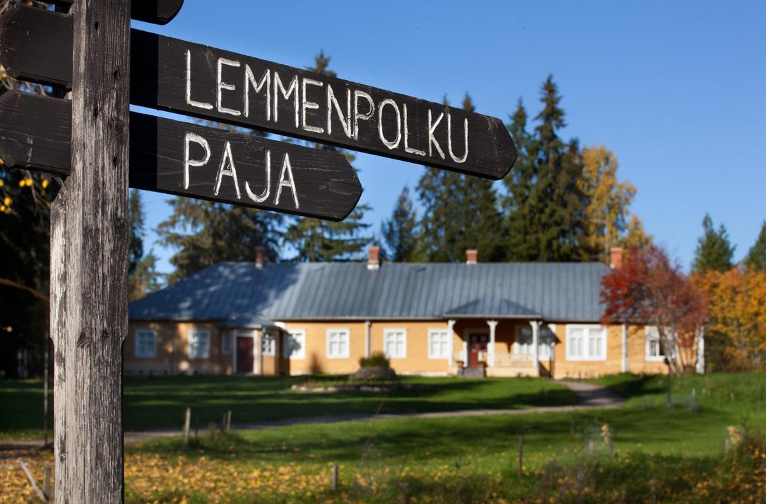 Musta puinen opasteviitta, jossa on valkoisella tekstit "LEMMENPOLKU" ja "PAJA". Taustalla näkyy epätarkkana Möhkön ruukkimuseon keltainen Pytinki-rakennus ja aurinkoinen nurmipiha.